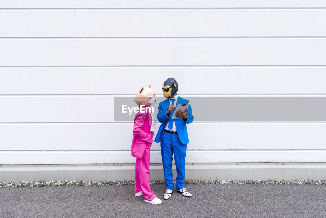 Man and woman wearing vibrant suits and animal masks talking over clipboard in front of white wall