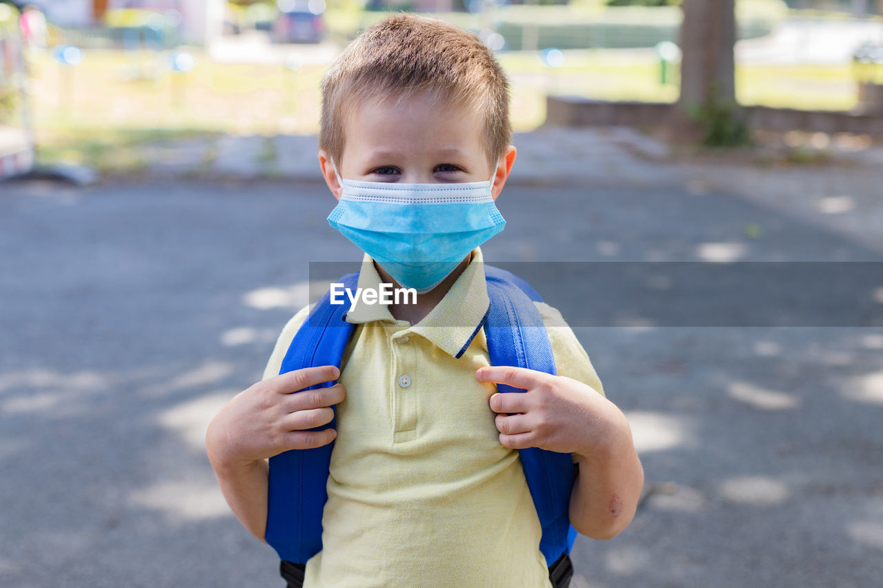 Boy wearing a protective mask with a backpack behind his back in the schoolyard 