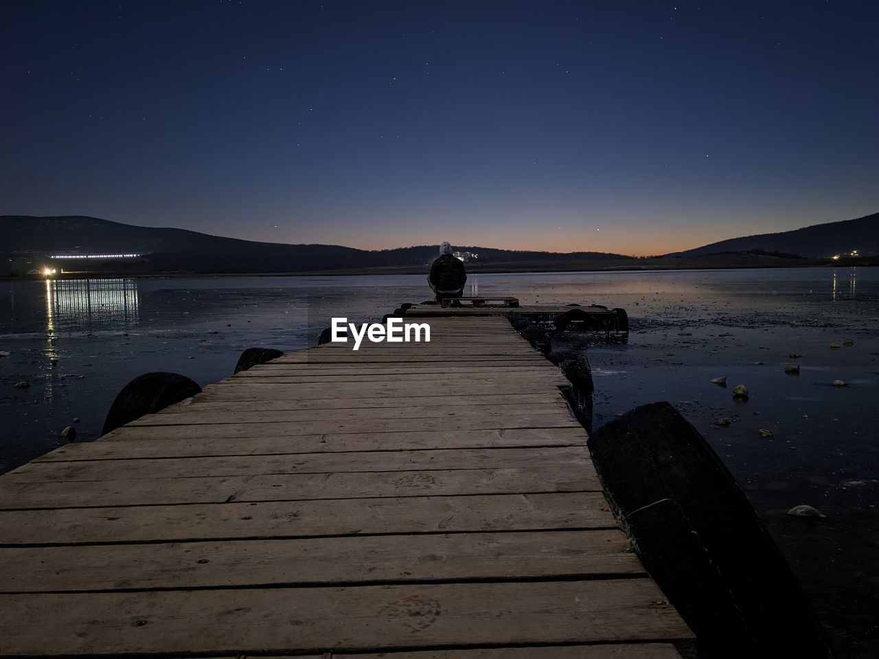 MAN SITTING ON PIER BY LAKE AGAINST SKY AT NIGHT