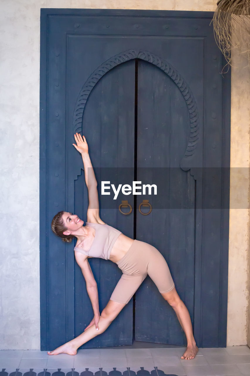 A slender woman in a beige top and breeches stands at a  blue wooden oriental door in a yoga pose