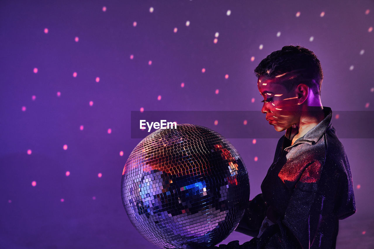 Side view of trendy young african american female with makeup and glitter ball in violet light