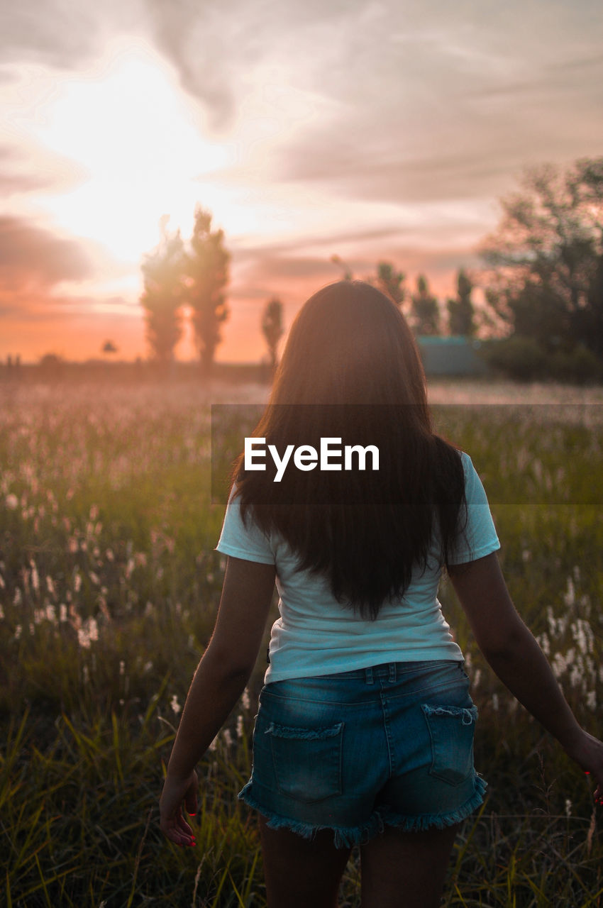 REAR VIEW OF WOMAN STANDING ON FIELD DURING SUNSET