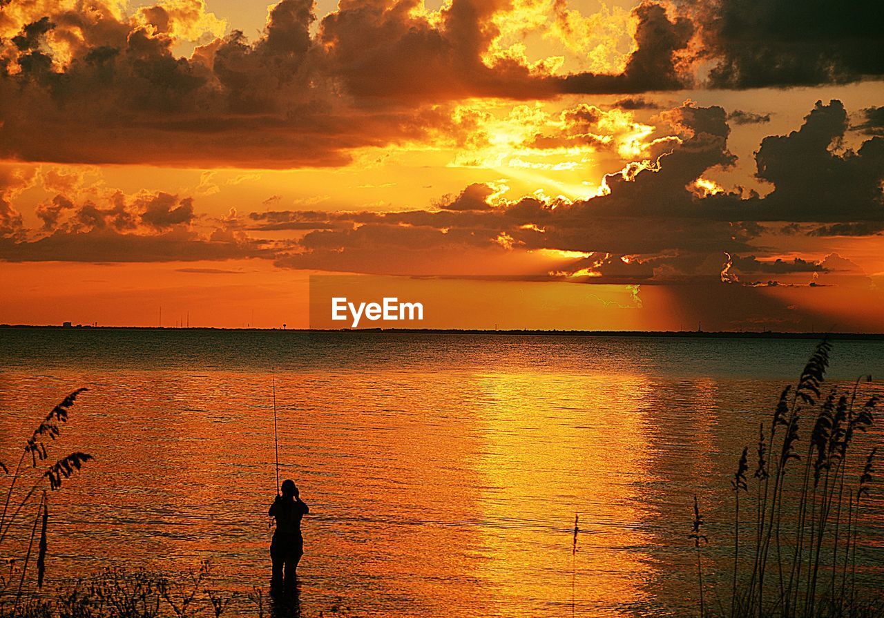 SILHOUETTE MAN STANDING IN SEA AGAINST ORANGE SKY