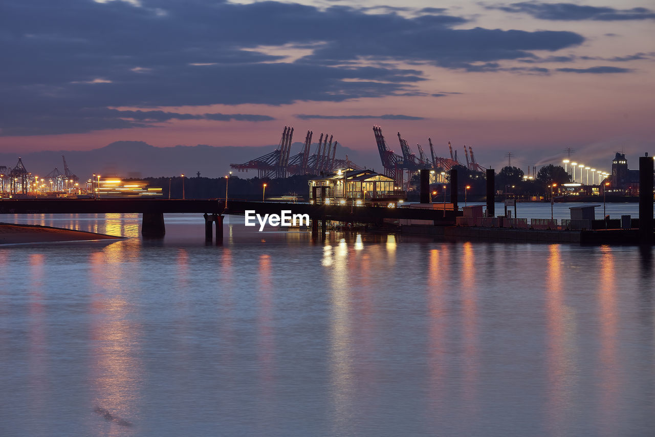 Illuminated pier by river against sky during sunset