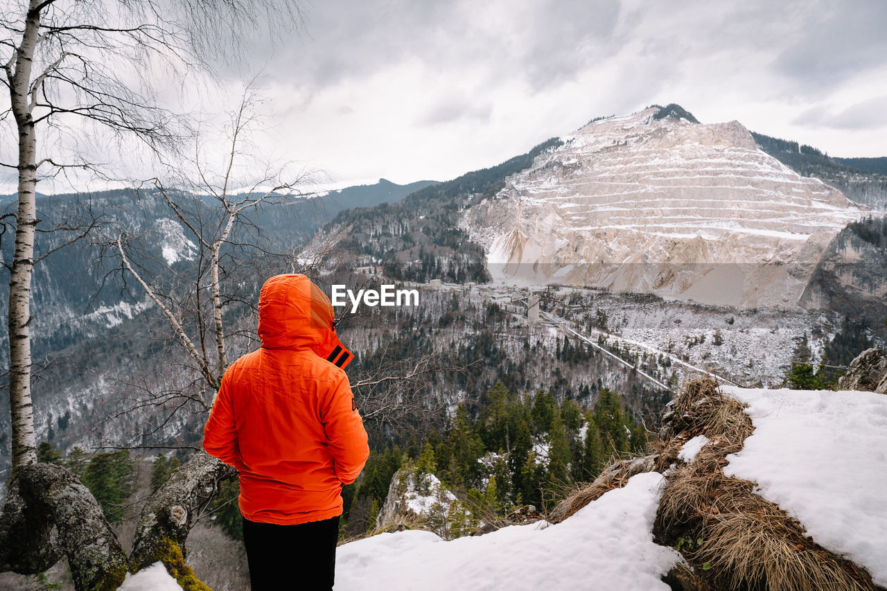 Rear view of person in orange jacket looking at snowcapped mountain during winter