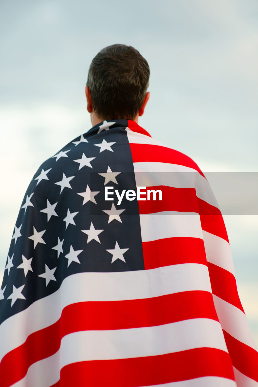 Rear view of man covered with american flag against sky