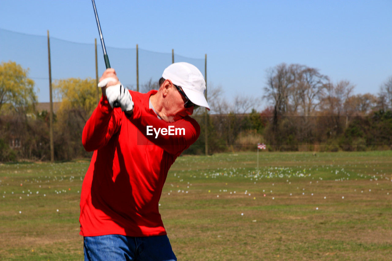 golf, sports, one person, recreation, adult, men, clothing, nature, leisure activity, activity, grass, holding, sky, shooting sport, day, red, motion, standing, golfer, plant, hat, three quarter length, golf course, outdoors, player