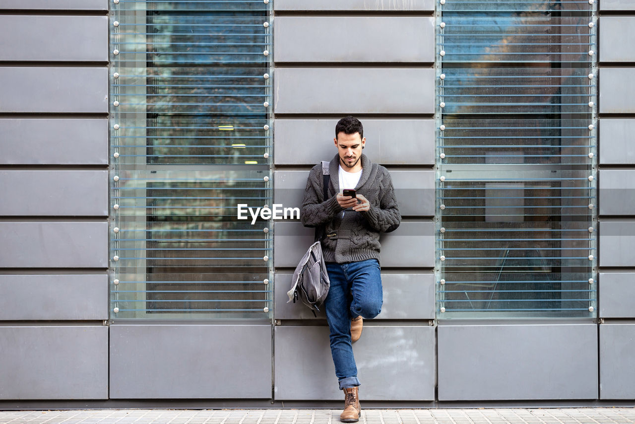 Front view of a bearded man using phone leaning on office building wall