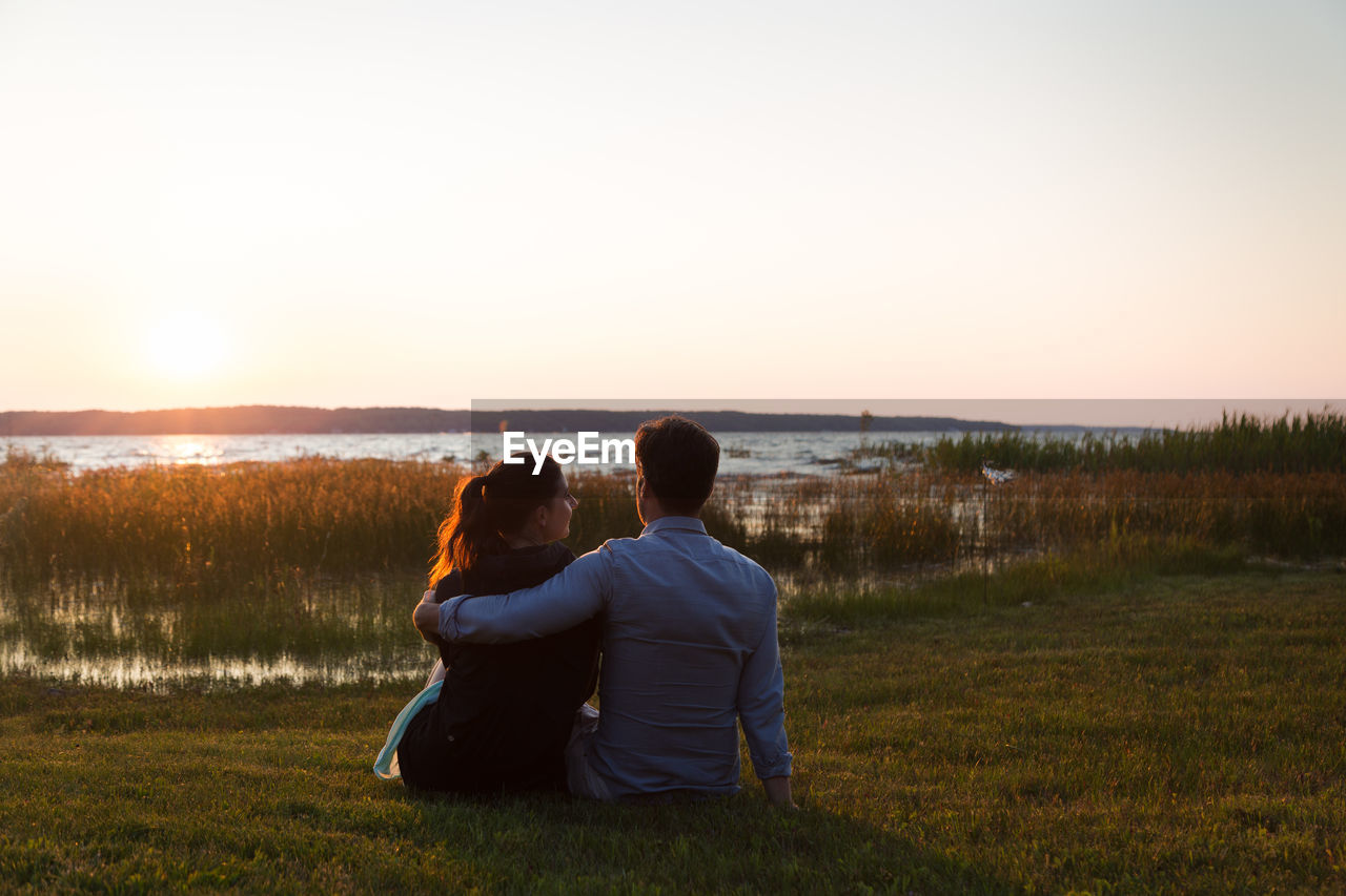 Rear view of couple sitting on grass against sky during sunset
