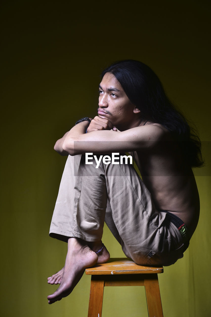 Side view of young man looking away while sitting on stool