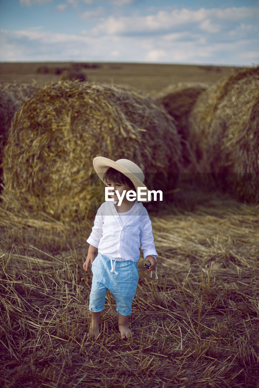 Boy a child in a straw hat and blue pants stands in a mowed field with stacks in the summer