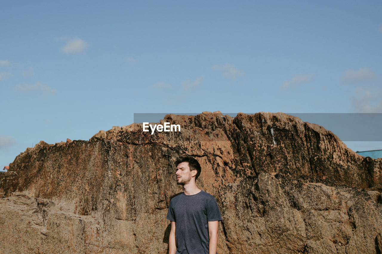 Young man standing by rock formation against sky