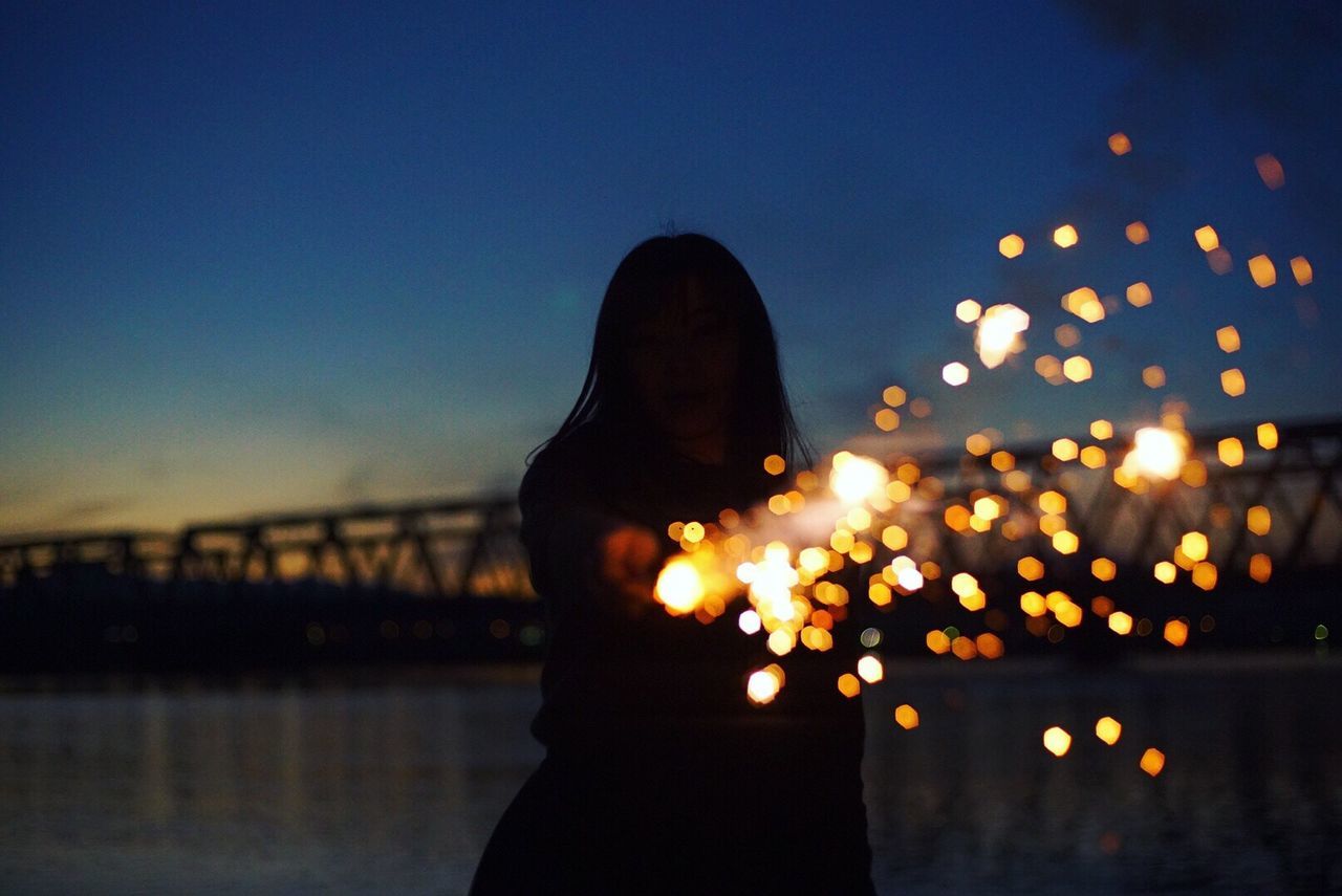 Silhouette woman with light painting by river against sky at dusk