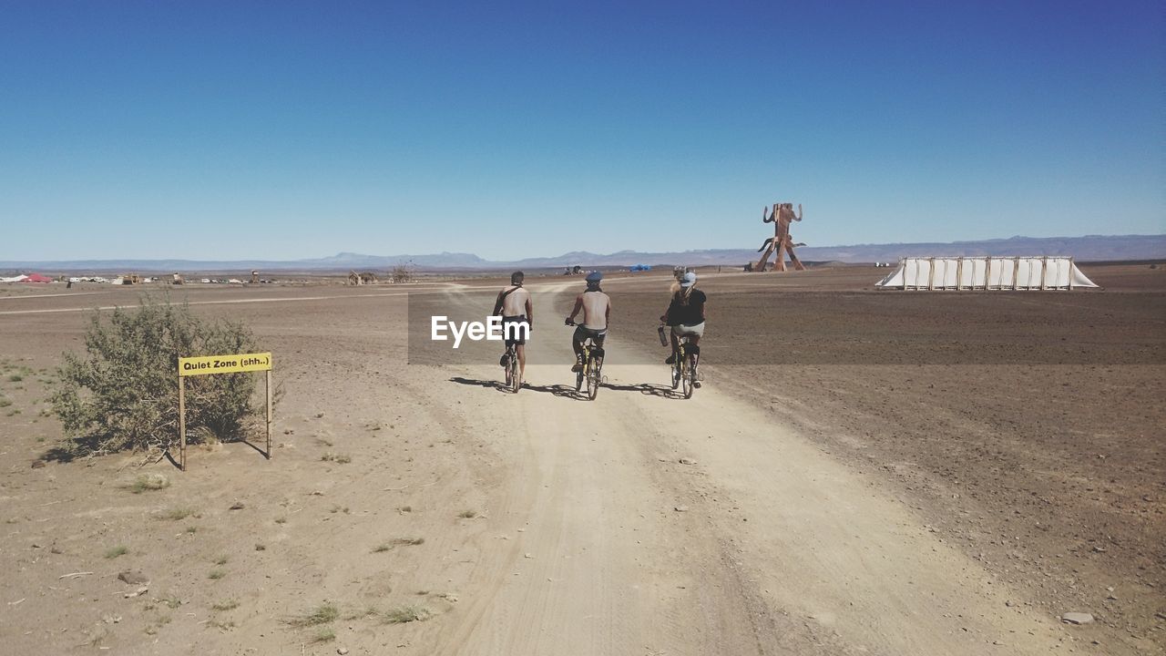 Rear view of men and woman cycling on dirt road against clear sky