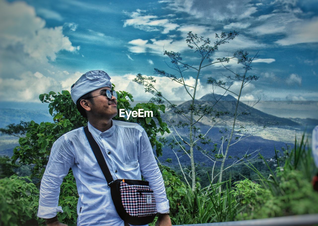 MAN STANDING BY TREE AGAINST SKY