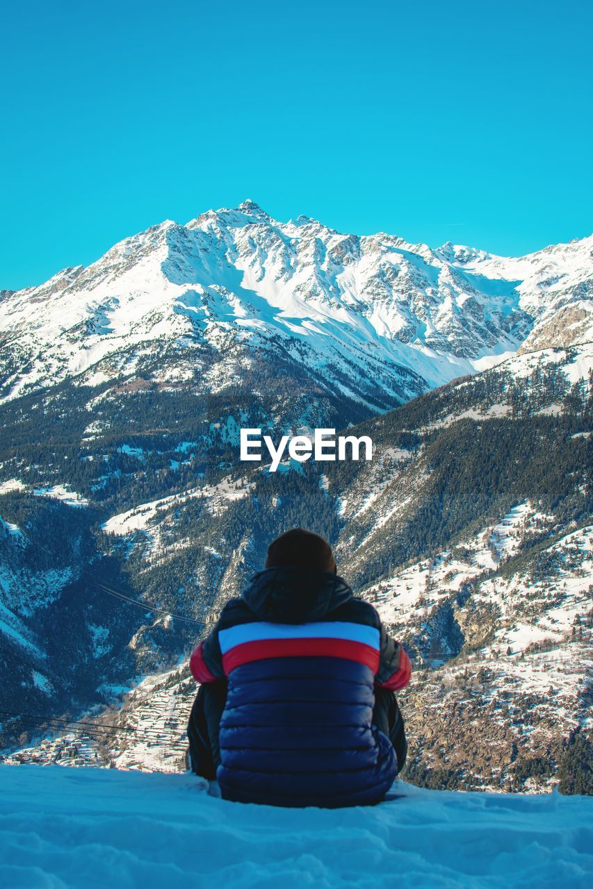 Rear view of boy sitting on snow covered land against mountains and sky