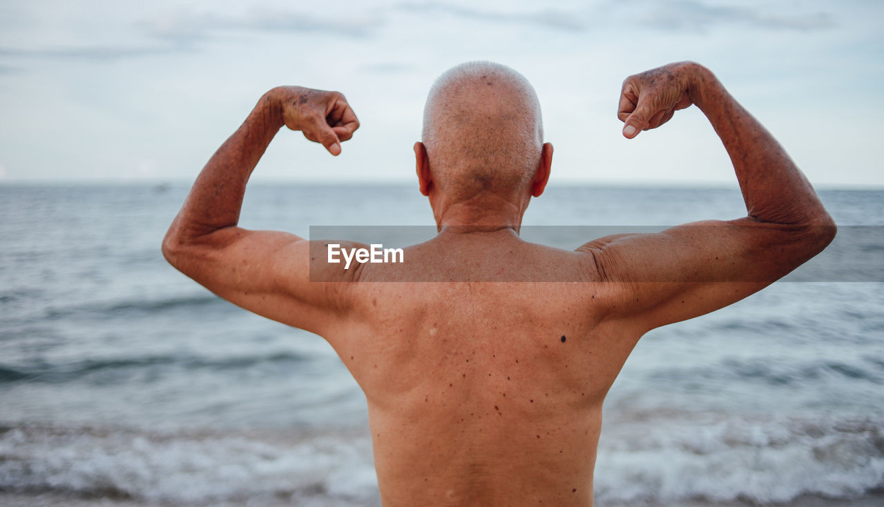 Rear view of shirtless senior man flexing muscles while standing at beach