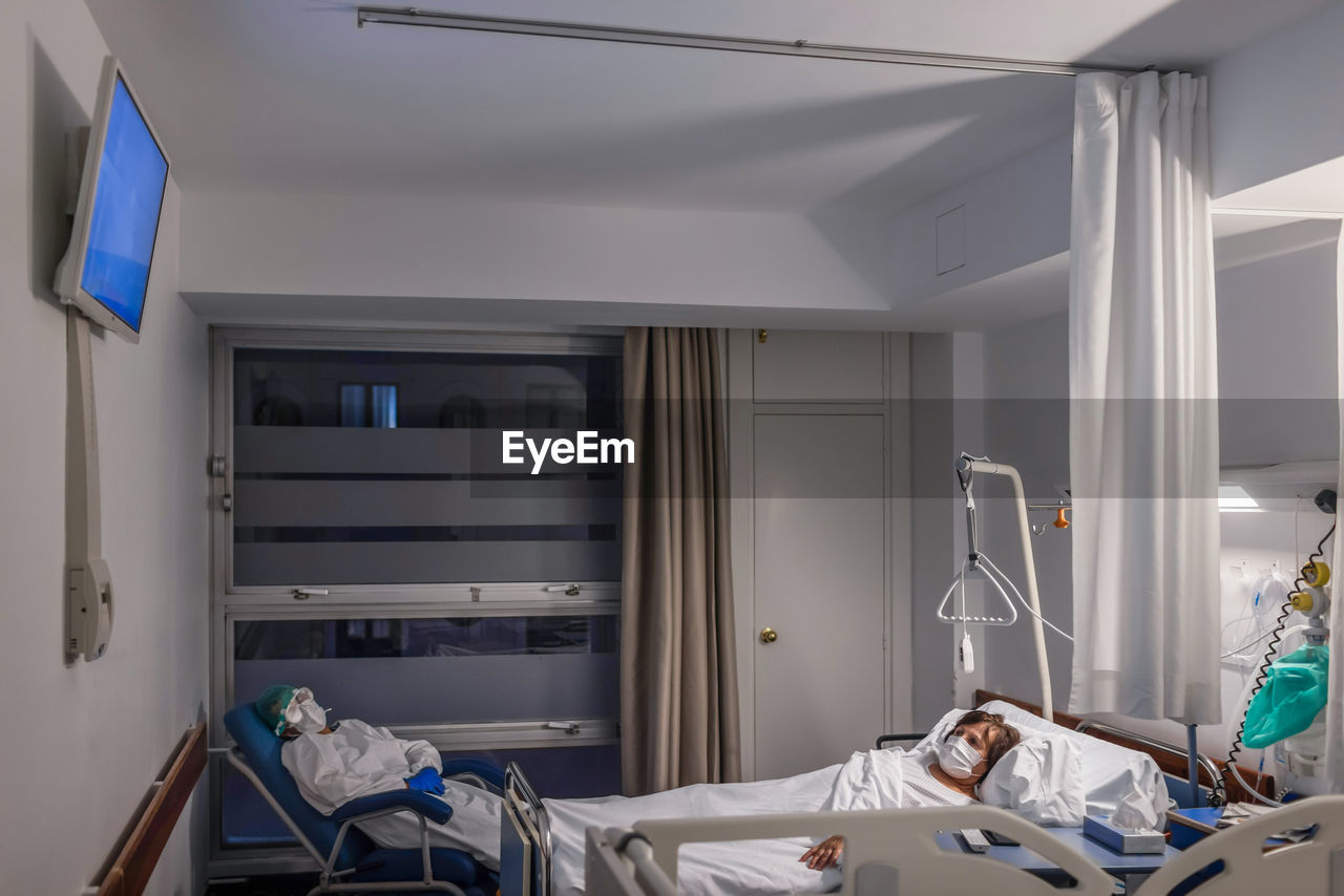 Side view of elderly female patient in medical mask lying on bed in hospital ward with visitor male wearing protective uniform sitting in armchair