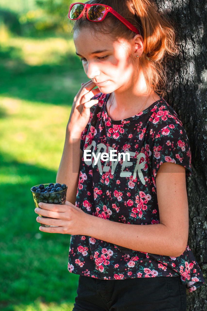 Thoughtful teenage holding blueberries in glass while standing in garden