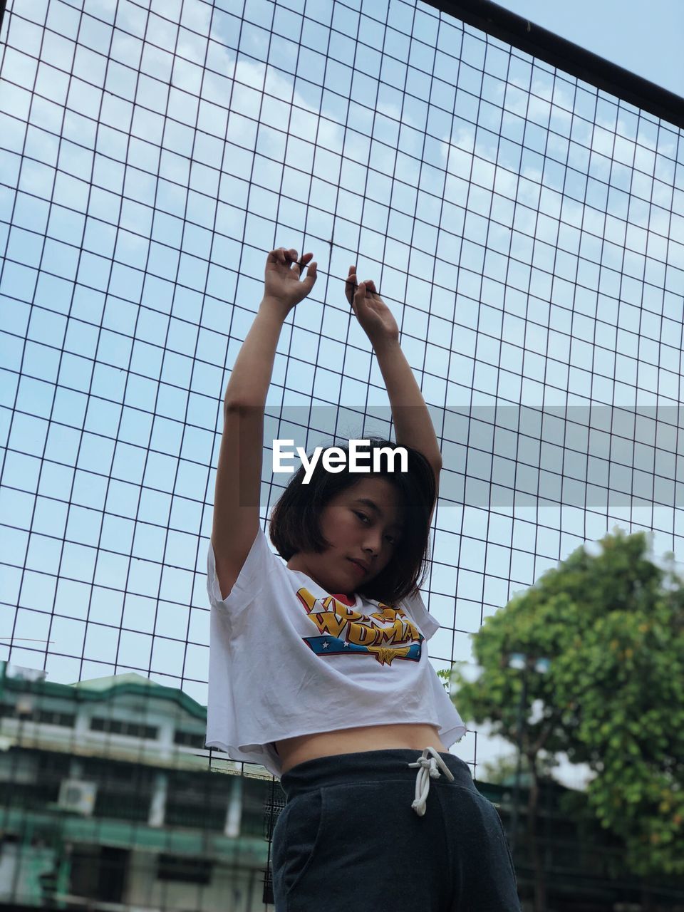 Portrait of young woman with arms raised standing against fence