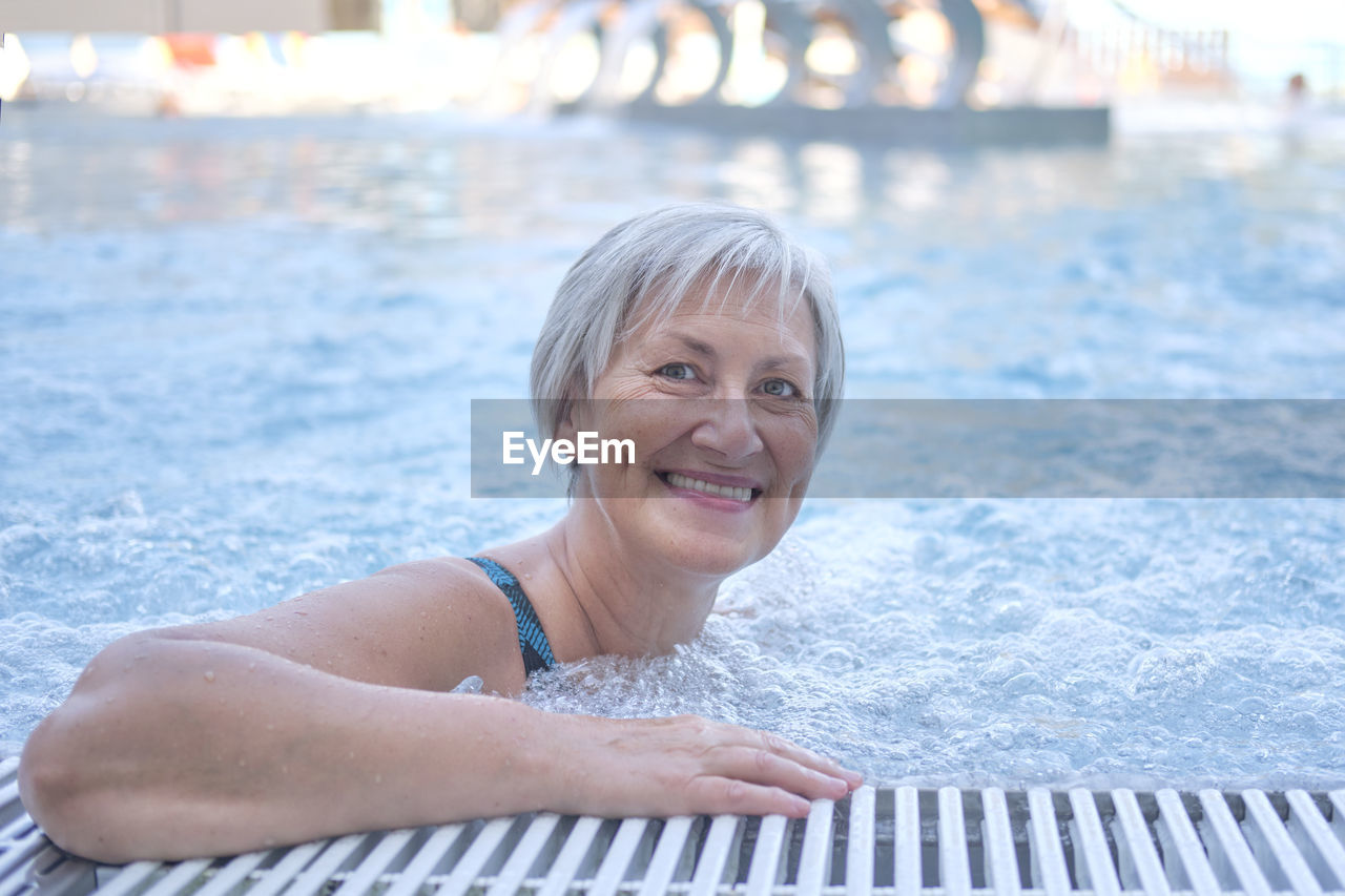 Happy senior woman with gray hair looking at camera in outdoor thermal pool with hydromassage. 