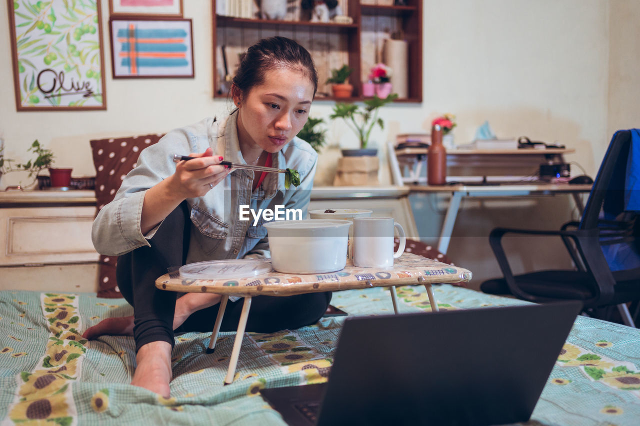 Focused asian female sitting on bed and eating food with chopsticks while watching movie with interest on laptop and chilling at weekend