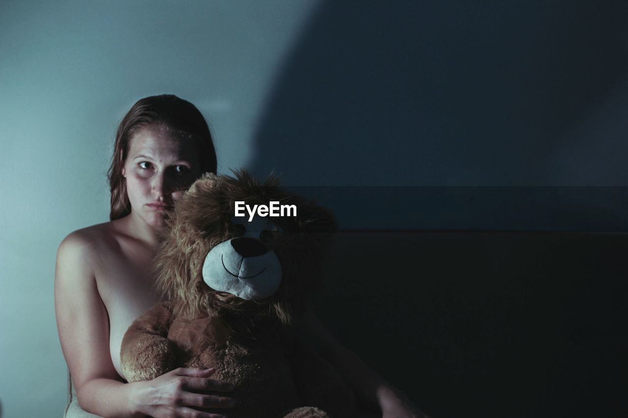 Portrait of naked woman holding teddy bear while sitting darkroom