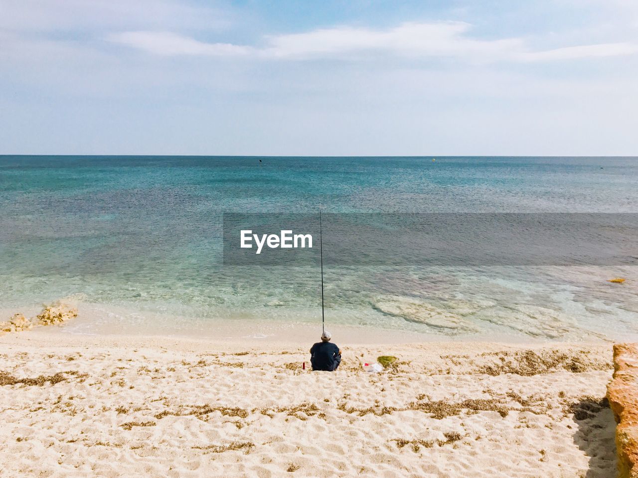 Rear view of man fishing while sitting on shore at beach against sky