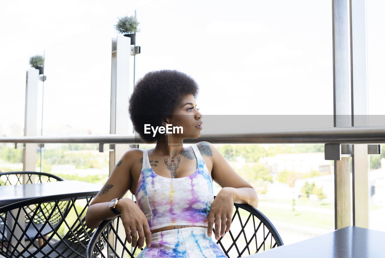 Calm black female with afro hairstyle resting in armchair of outdoors cafe and waiting for order while looking away