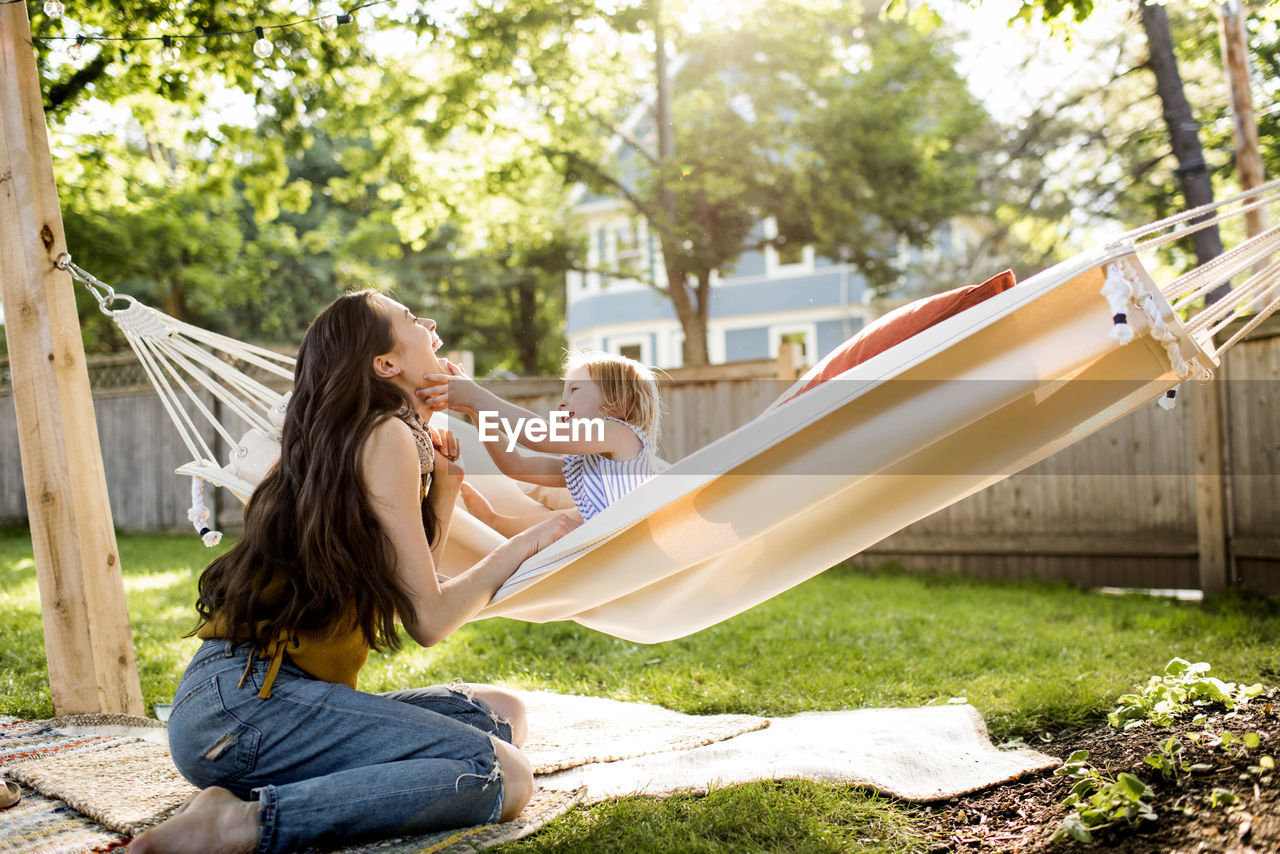 Happy mother playing with daughter sitting in hammock at yard