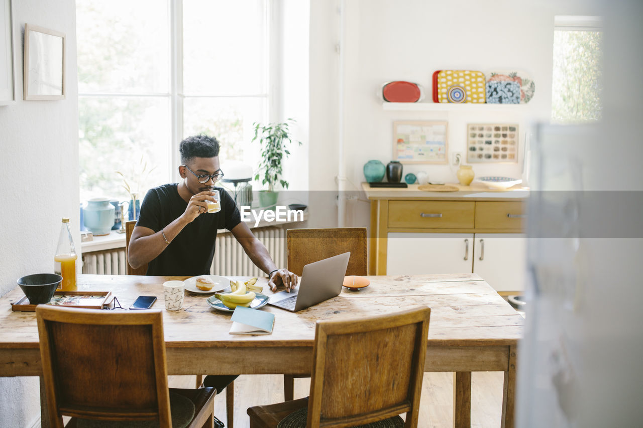 Young man drinking juice while using laptop at table in house