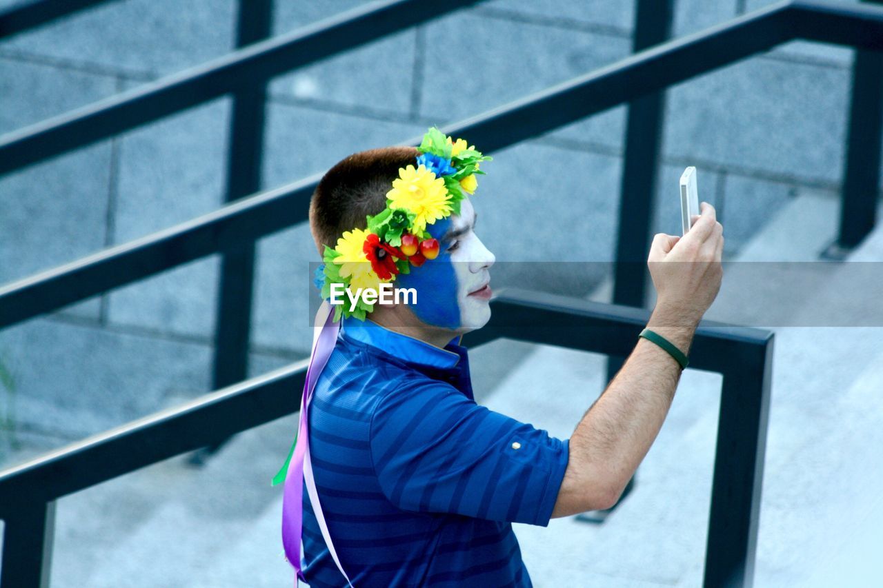Side view of man with painted face wearing flowers taking selfie through mobile phone