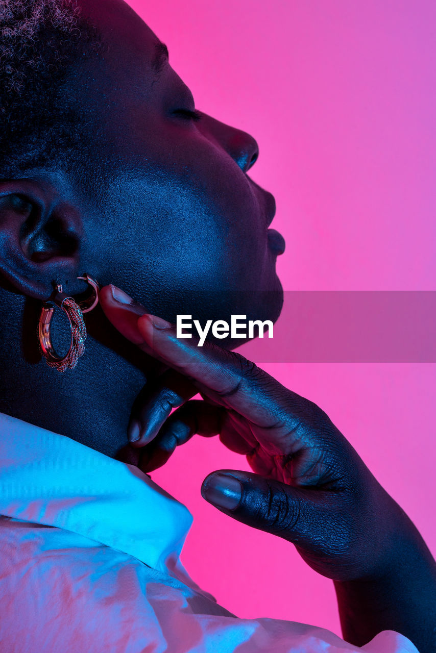 Side view of delicate african american female model in trendy earrings touching neck in studio with neon light on pink background