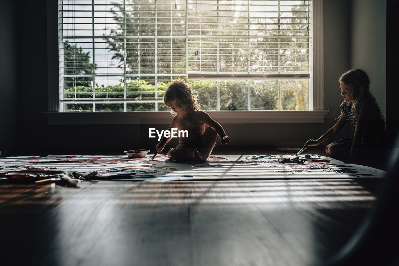 Sisters painting while sitting on floor against window at home