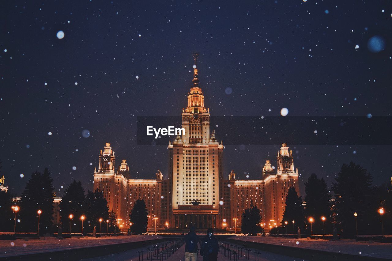 Rear view of young couple standing near illuminated historic buildings during snowfall at night