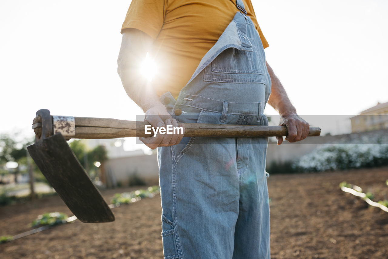 Senior man in denim holding shabby hoe tool and standing on cultivated land