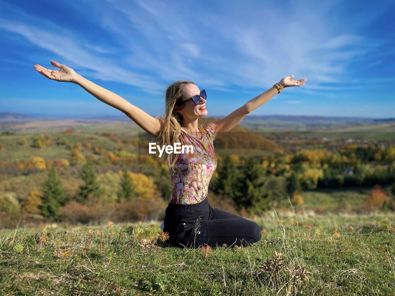 Young woman with her arms outstretched sitting down on the grass admiring the beautiful fall scenery