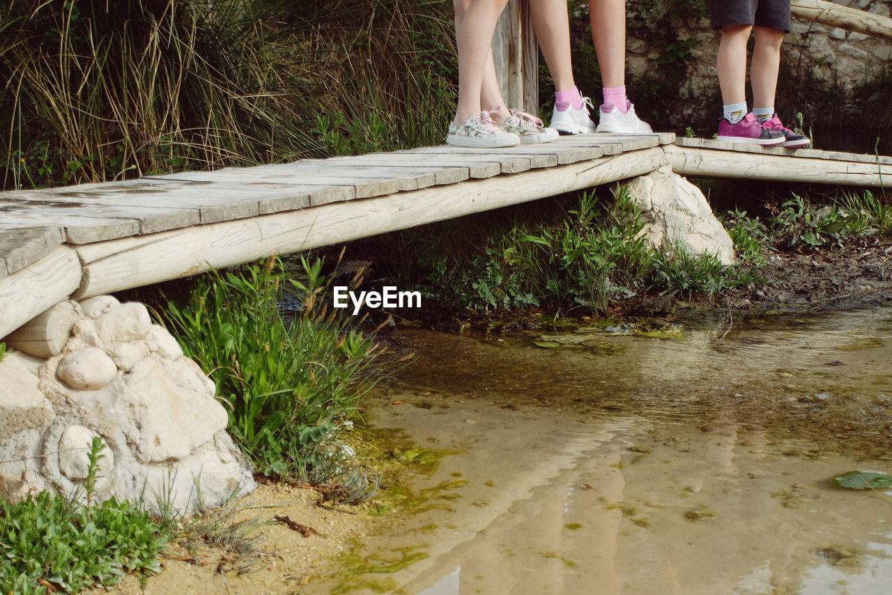 Low section of children standing on wooden walkway over stream