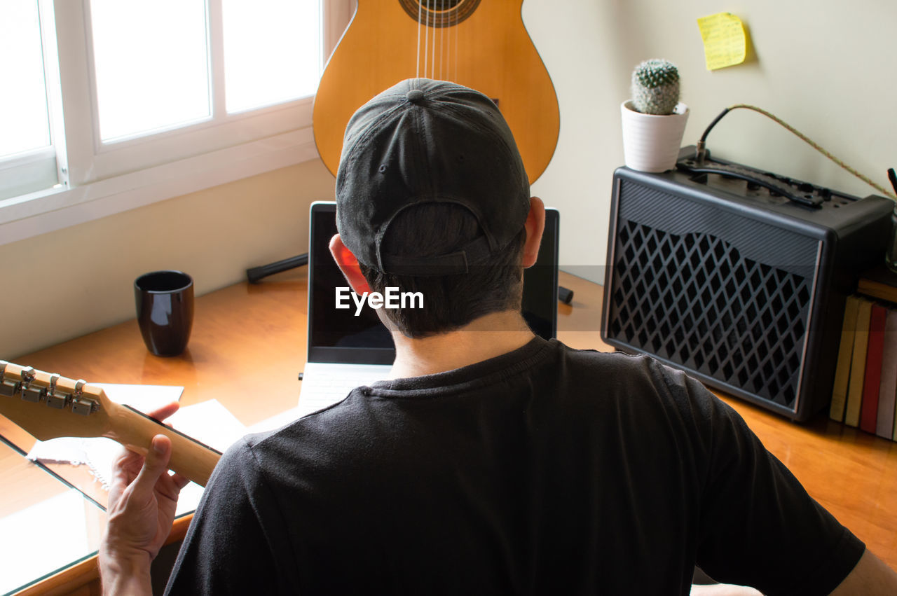 Rear view of man playing guitar while using laptop at home
