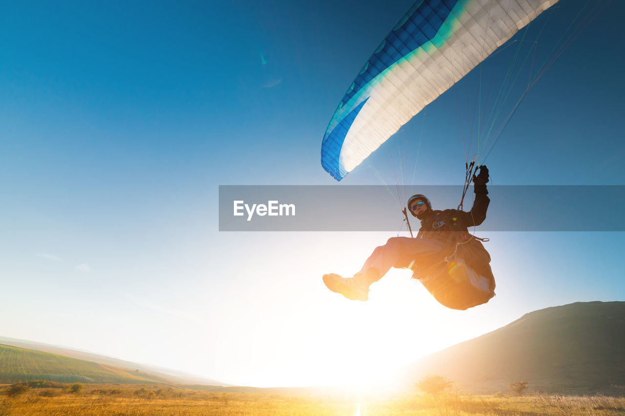 A paraglider takes off from a mountainside with a blue and white canopy and the sun behind. a