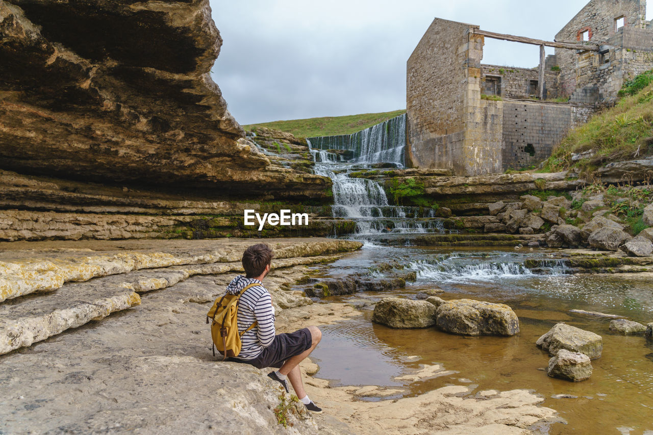 REAR VIEW OF MAN SITTING ON ROCK BY WATER