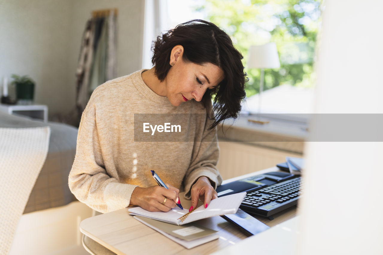 Mature businesswoman writing in note pad on table at home