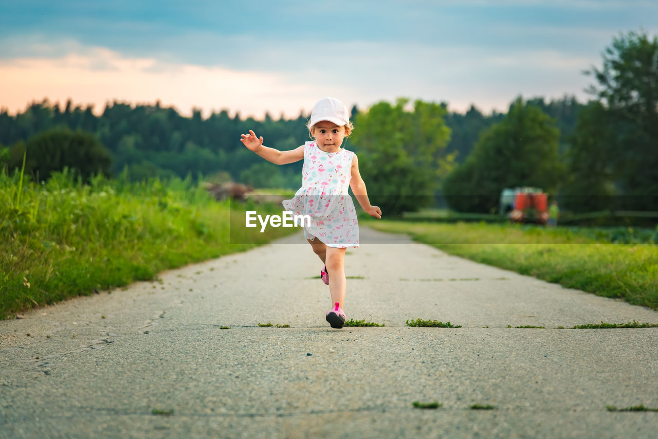 Baby girl with cap outdoors running on a rural road leading from forest.