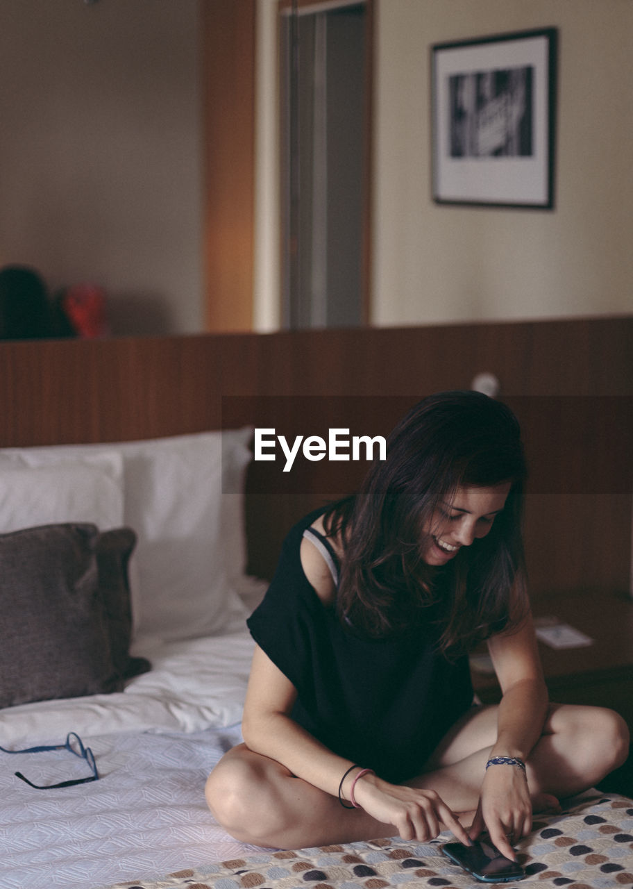 A girl sitting in the bed, reading emails using a smartphone with glasses off