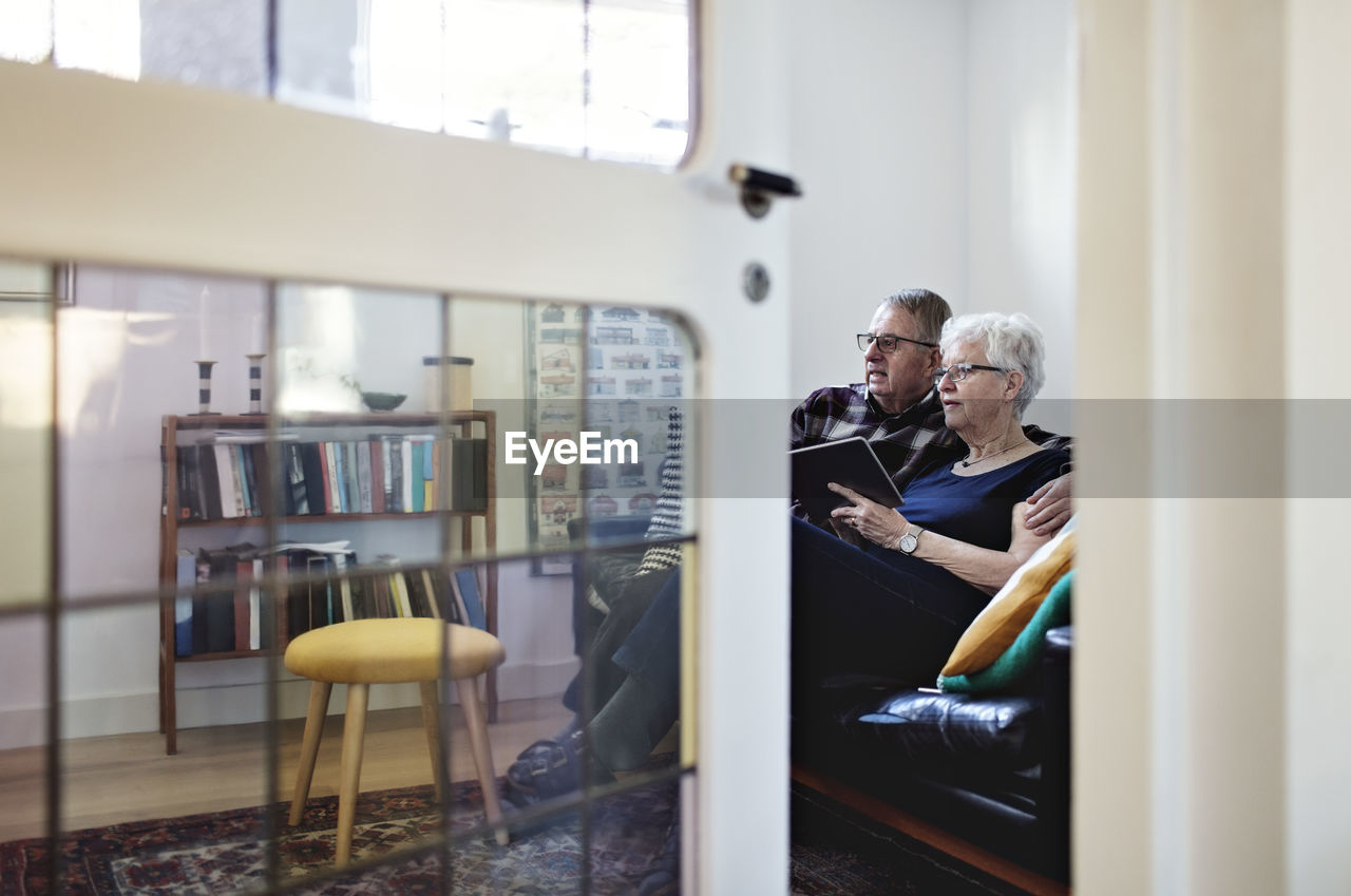 Full length of senior couple sitting on sofa with digital tablet seen from ajar door