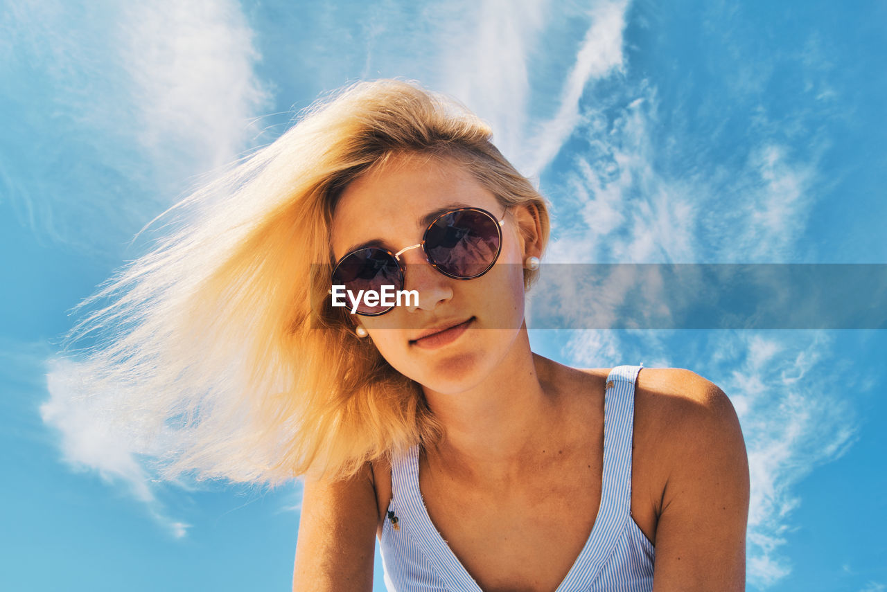 Low angle portrait of young woman wearing sunglasses while bending against sky