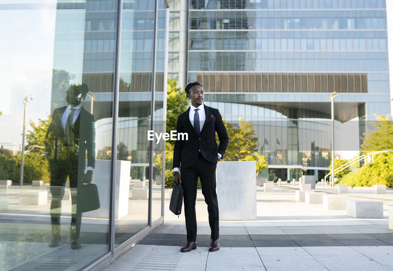 Serious african american male entrepreneur wearing elegant suit walking along glass office building in city center and looking away