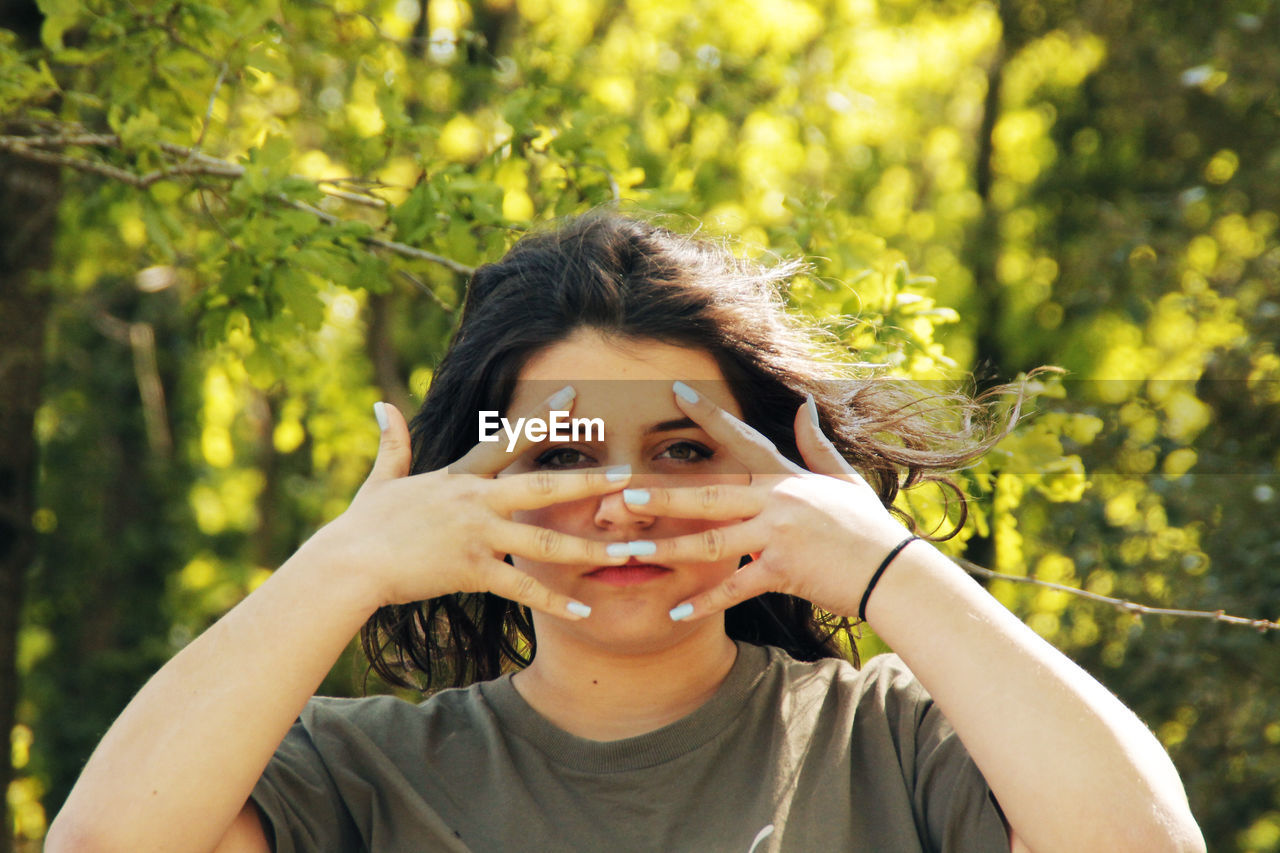 Portrait of young woman peeking through fingers against trees