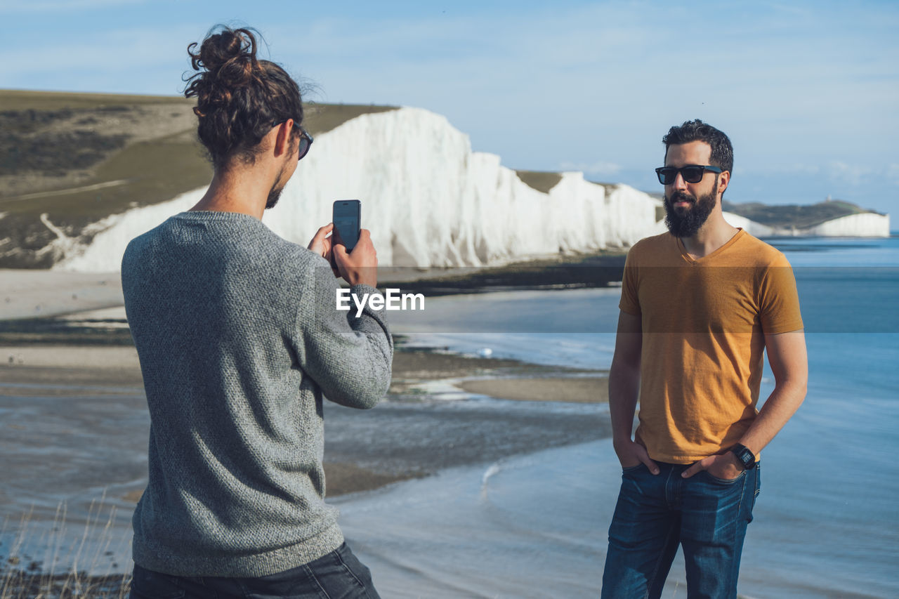 Back view of unrecognizable trendy guy taking photo of male bearded ethnic friend while spending holidays together in front of chalk cliff of seven sisters