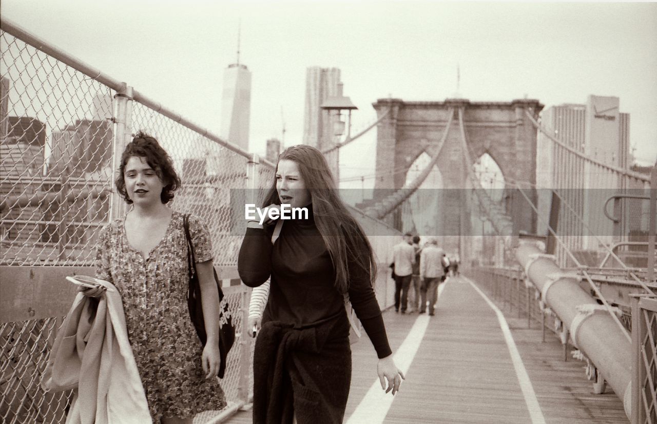 FULL LENGTH OF SMILING YOUNG WOMAN STANDING ON BRIDGE IN CITY AGAINST SKY